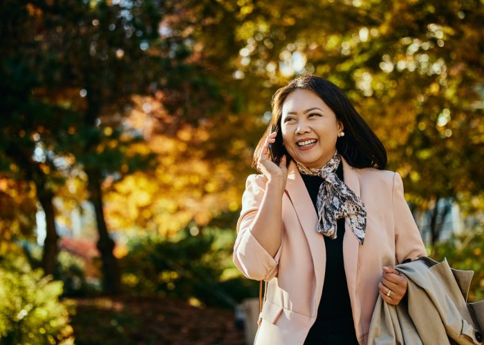 Happy Asian businesswoman communicating on cell phone while walking through city park.