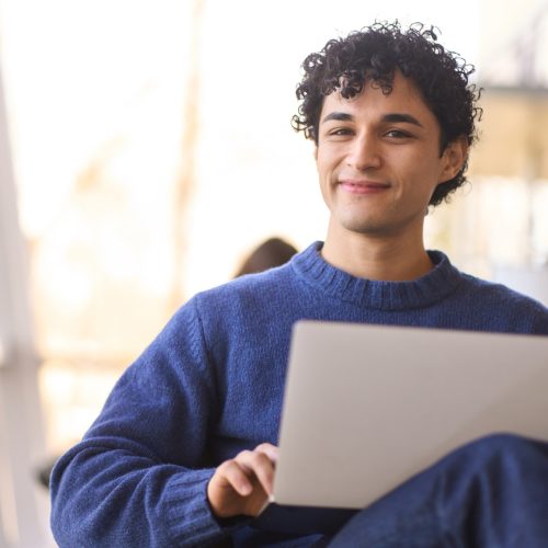 Portrait of a handsome smiling positive young Latin American man with laptop, looking confidently at camera. Businessman telecommuting in a modern office interior. People. Online business concept